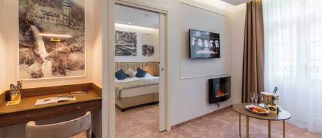 A hotel room with a bed, a desk with a chair, a television, a fireplace, and a dining table with a plate of fruit.