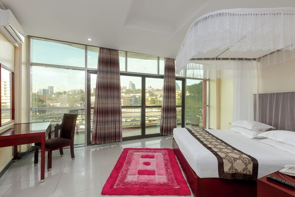 A hotel room with a large bed, a desk, and a view of the cityscape through large windows.