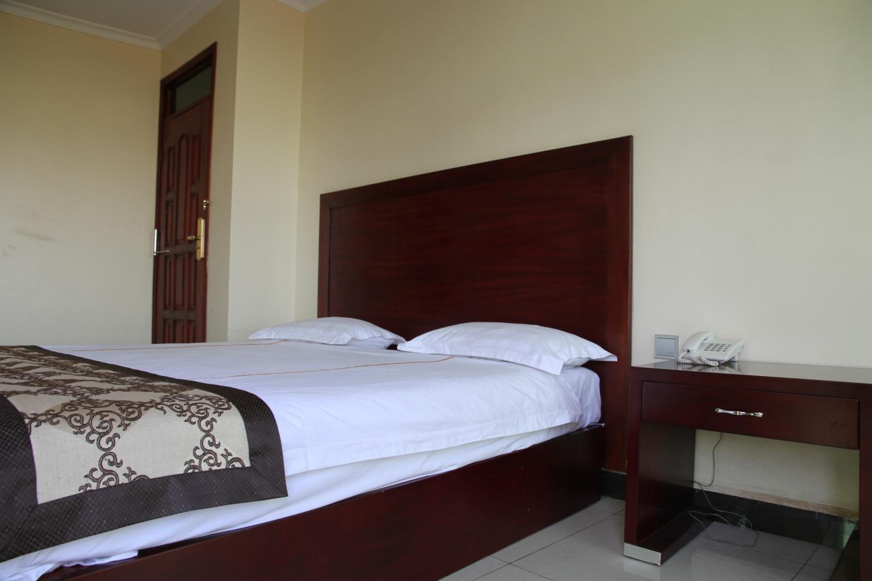 Una cama bien tendida con cabecera de madera, una mesita de noche con teléfono y una puerta al fondo.