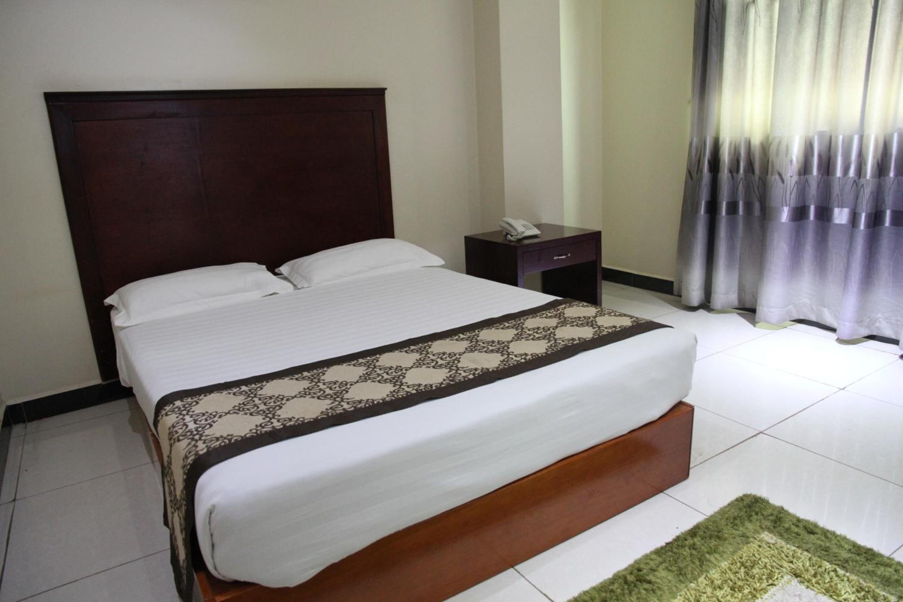 Uma cama bem arrumada com colcha estampada, cabeceira de madeira, mesa de cabeceira com telefone e tapete verde sobre piso de azulejos.