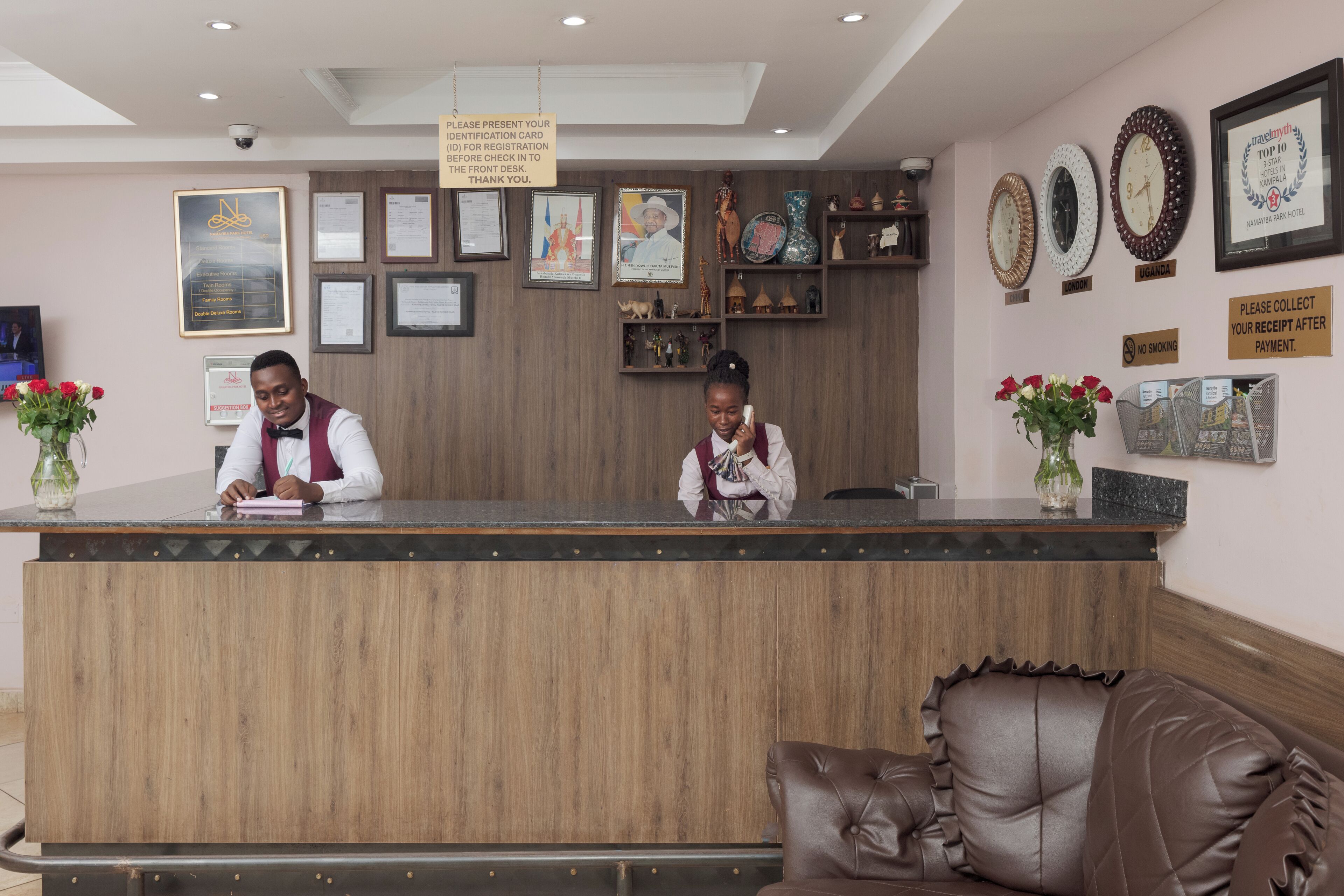 Un comptoir d’accueil d’hôtel avec deux employés, un comptoir en bois, des certificats encadrés et un mur orné de différentes plaques et d’un panneau « Interdiction de fumer ».