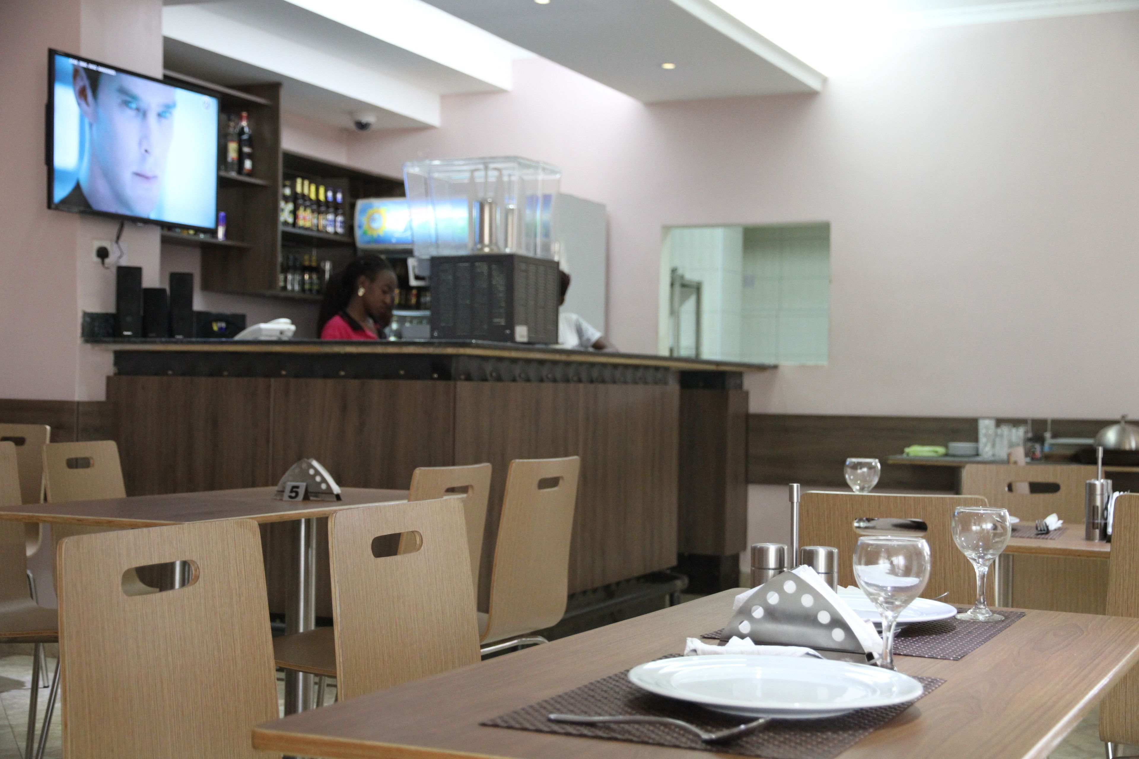 A restaurant with wooden tables and chairs, a bar area with a television, and a counter with a person behind it.