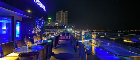 Un bar en la azotea con mobiliario de mimbre, vistas a una piscina y un paisaje urbano de fondo. Un letrero dice 'Sky Lounge'.
