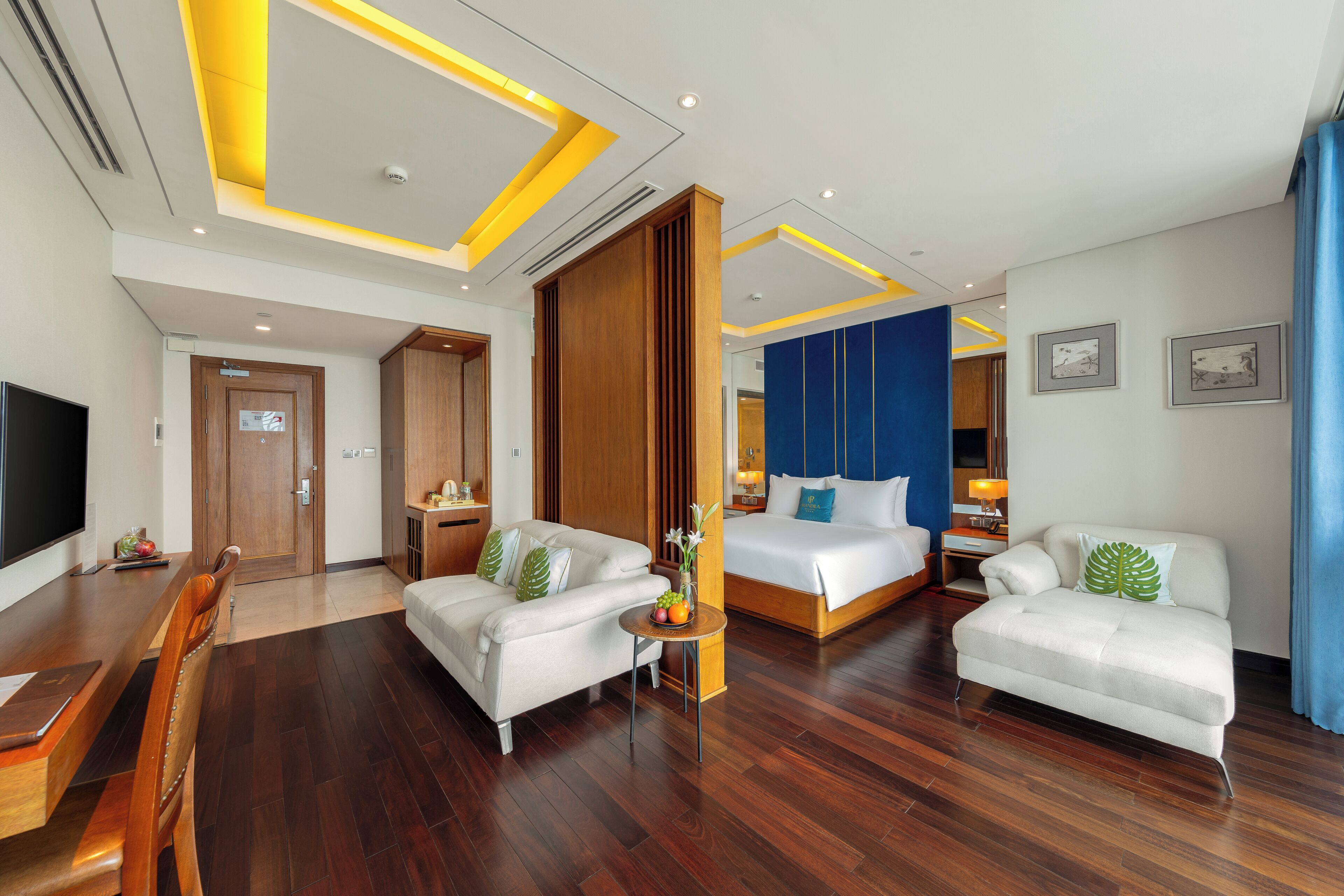 A modern hotel room with a wooden floor, a sofa, a bed, a TV, and a desk.