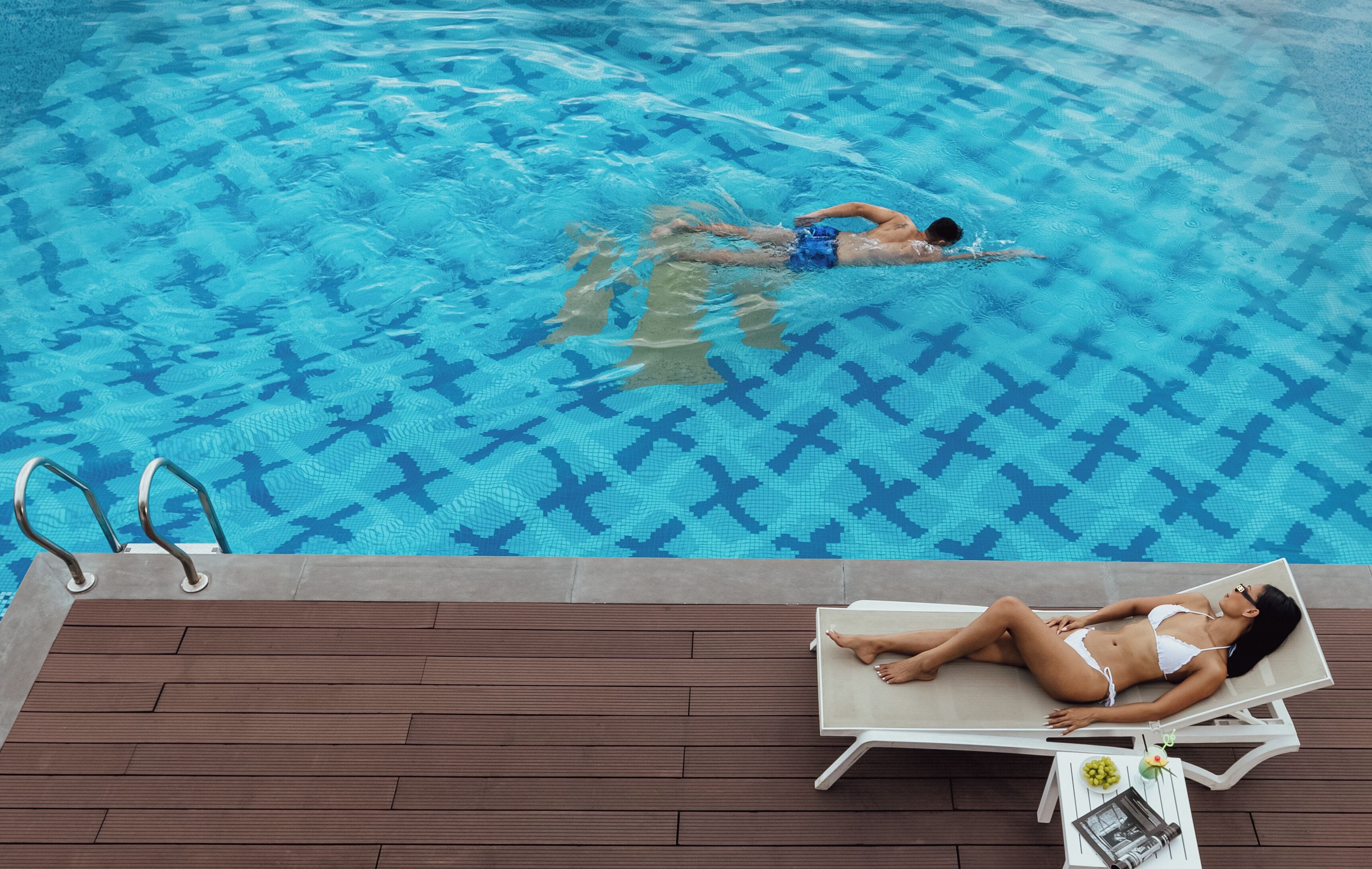 A swimming pool with a person swimming and another person lying on a lounge chair.