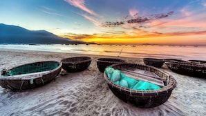 Una playa con varias embarcaciones tradicionales alineadas en la arena, un mar tranquilo y una puesta de sol al fondo.