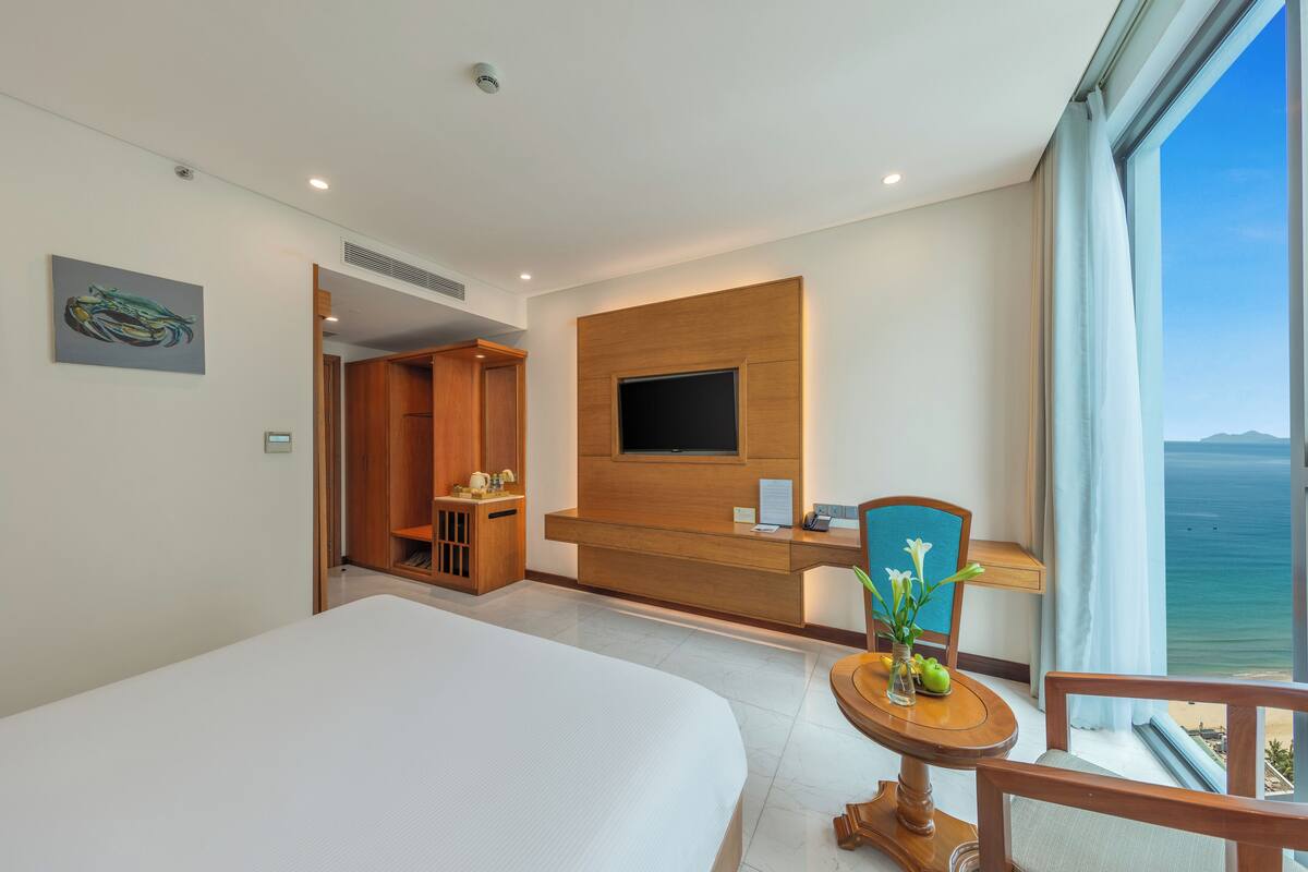 Een hotelkamer met een groot bed, een aan de muur bevestigde televisie, een bureau met een stoel, en uitzicht op de oceaan.