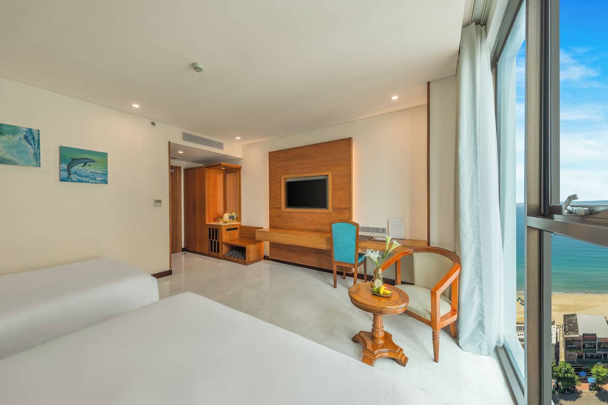 Hotelkamer met een bed, een bureau, een stoel, een tafeltje met een vaas en uitzicht op het strand.