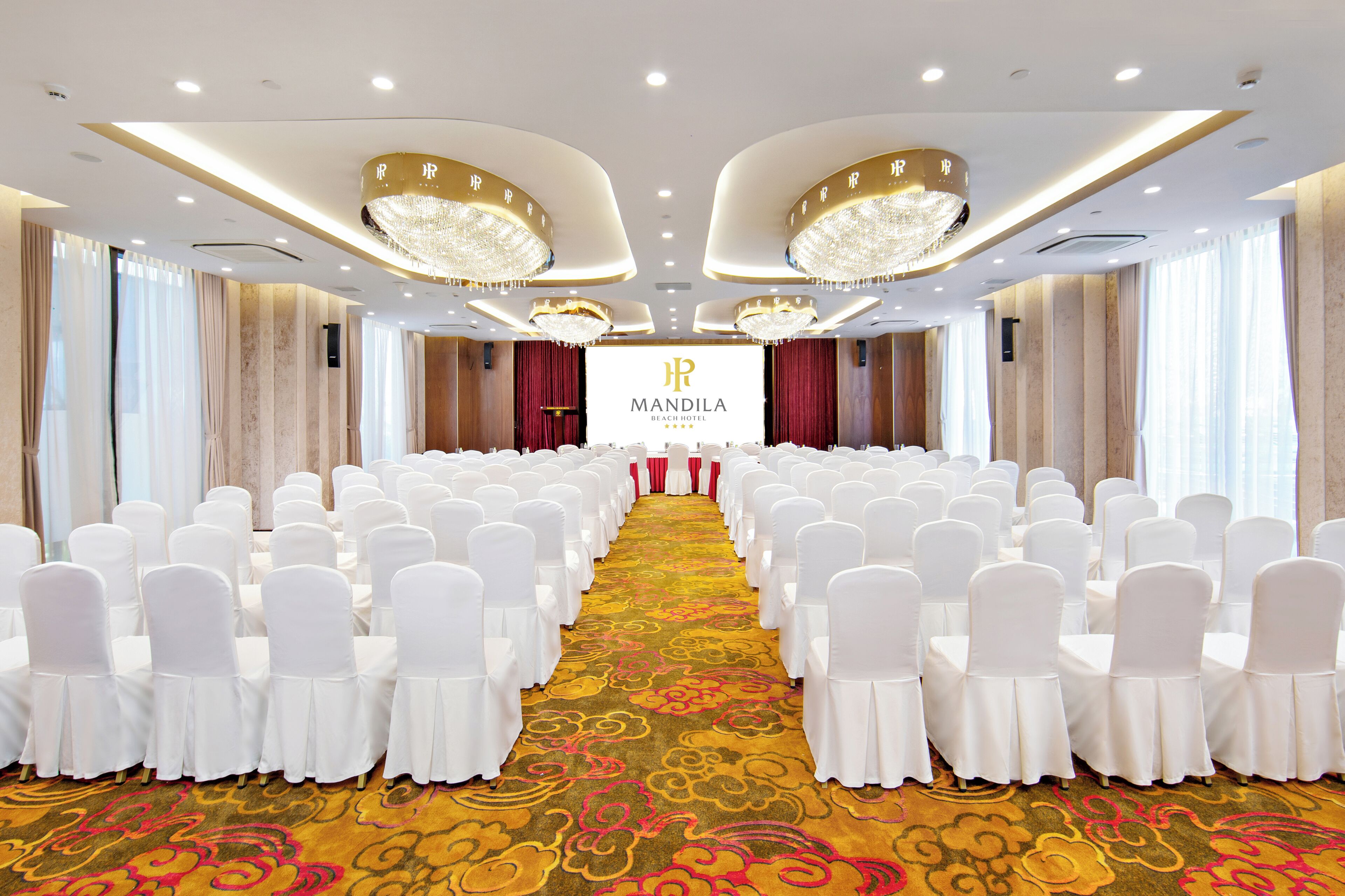 A conference room with rows of white chairs, a central stage with a banner, and chandeliers.