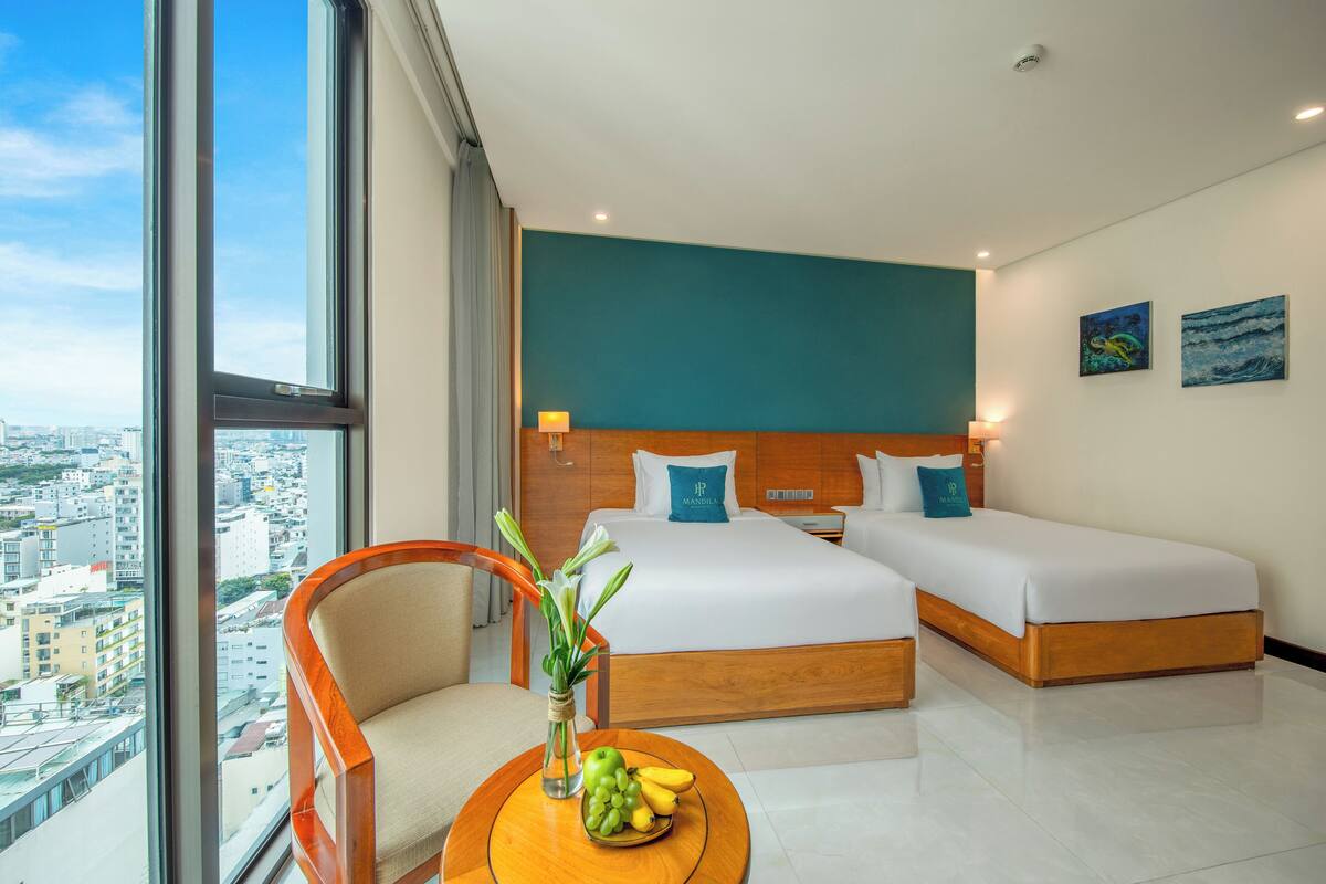 Hotelkamer met uitzicht op de stad, twee bedden, een houten hoofdbord, een ronde tafel met fruit en een stoel.