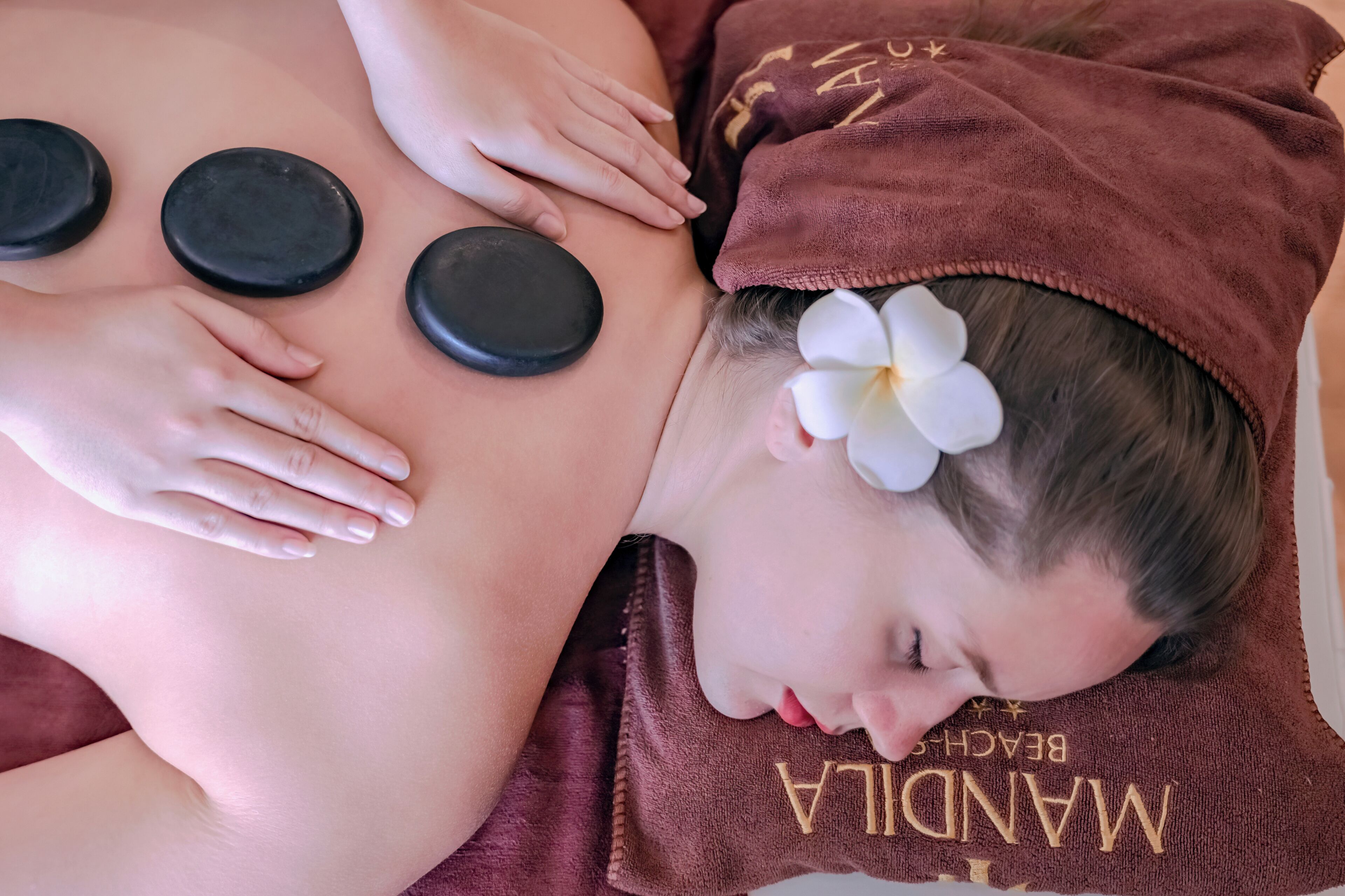 A person receiving a back massage with hot stones, lying on a towel with a flower accessory.