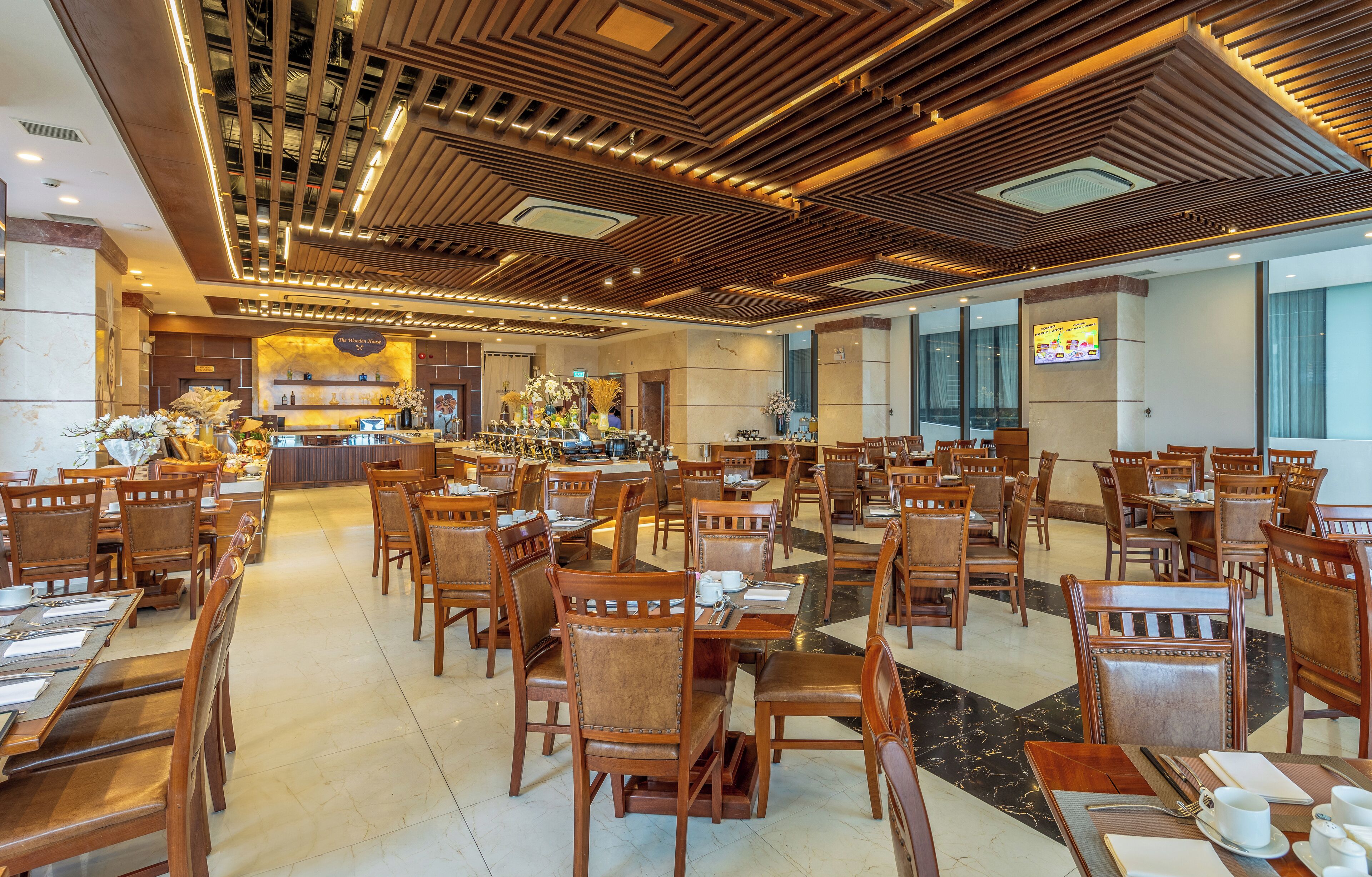 A spacious hotel lobby with wooden flooring, multiple dining tables and chairs, a reception area, and a large TV screen on the wall.