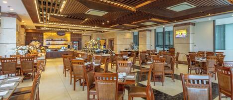 A spacious hotel lobby with wooden flooring, multiple dining tables and chairs, a reception area, and a large TV screen on the wall.