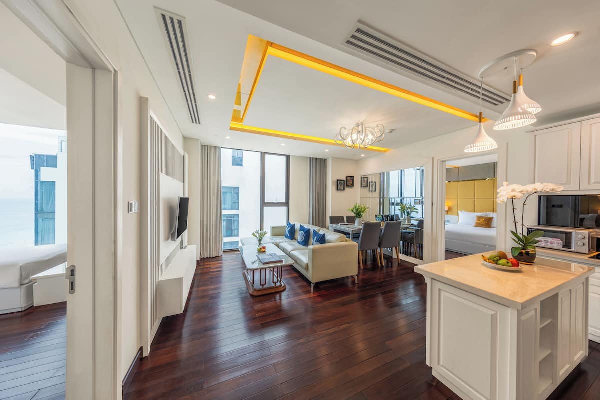 Een moderne woonkamer met een bank, salontafel en een eethoek. Er is een keuken met witte kasten en een groot eiland. De kamer heeft houten vloeren en grote ramen.