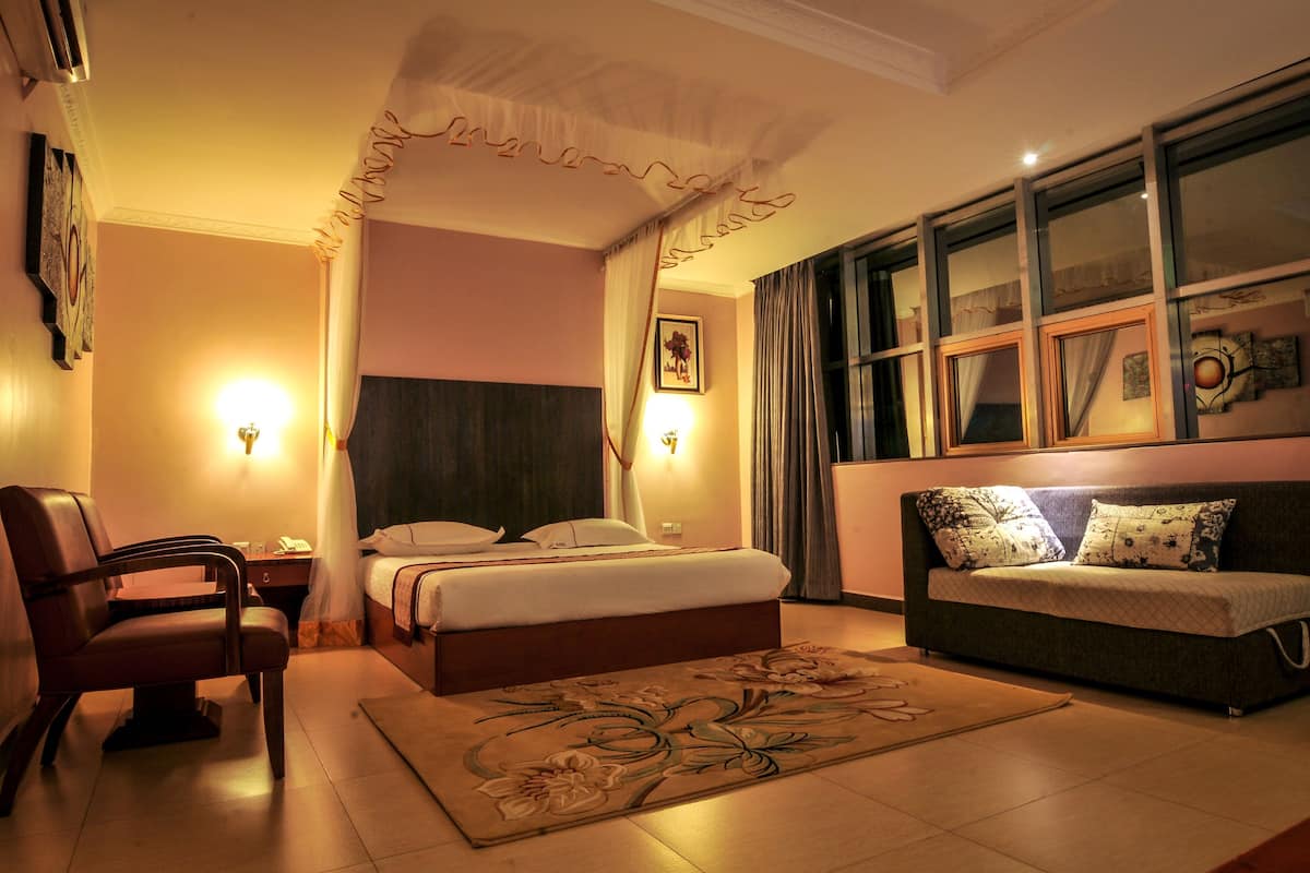 Una habitación de hotel con cama, sofá, sillón, ventana con cortinas e iluminación en la pared.