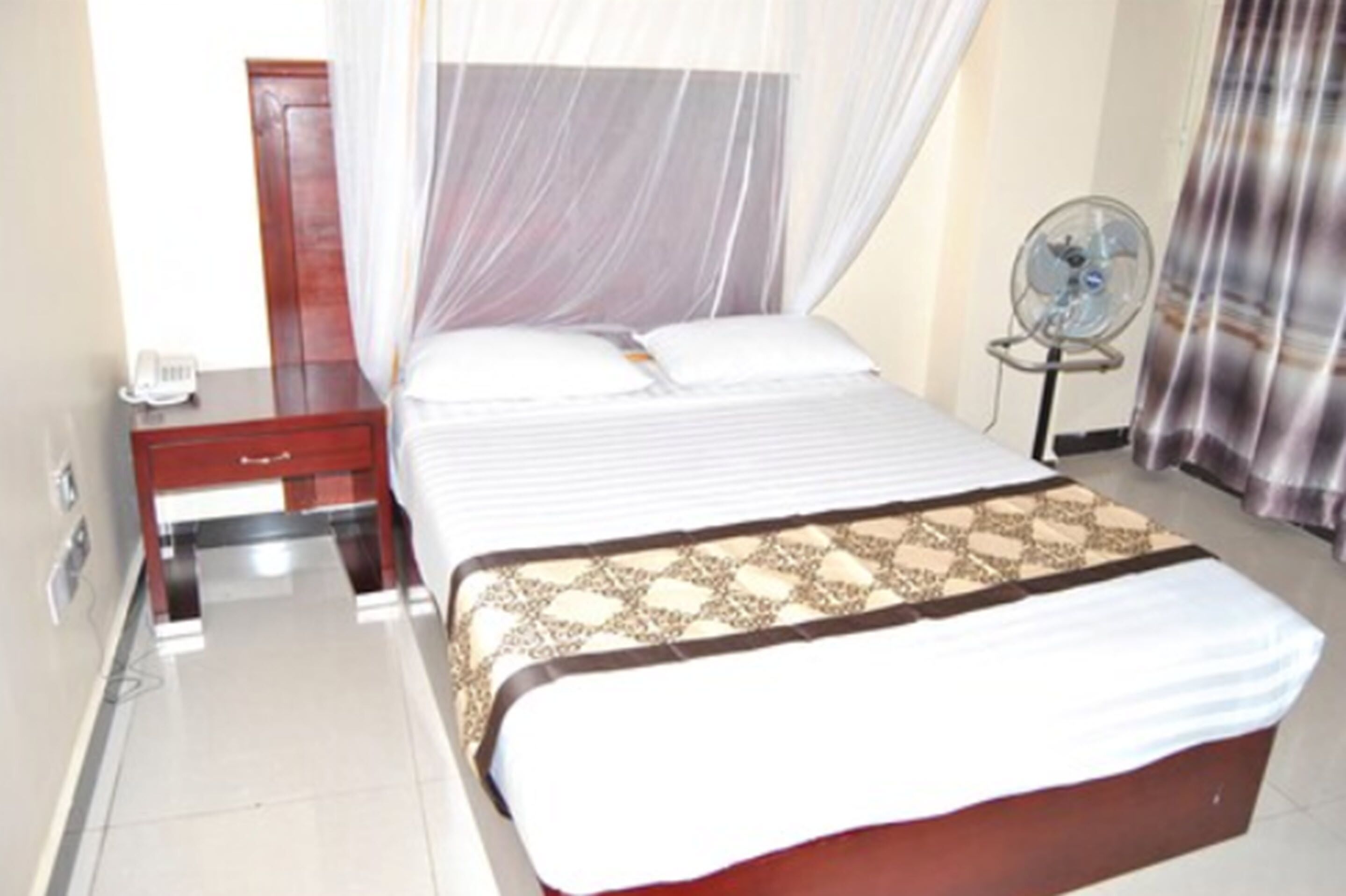 A hotel room with a bed, a bedside table with a phone, a bedside fan, and a curtain.