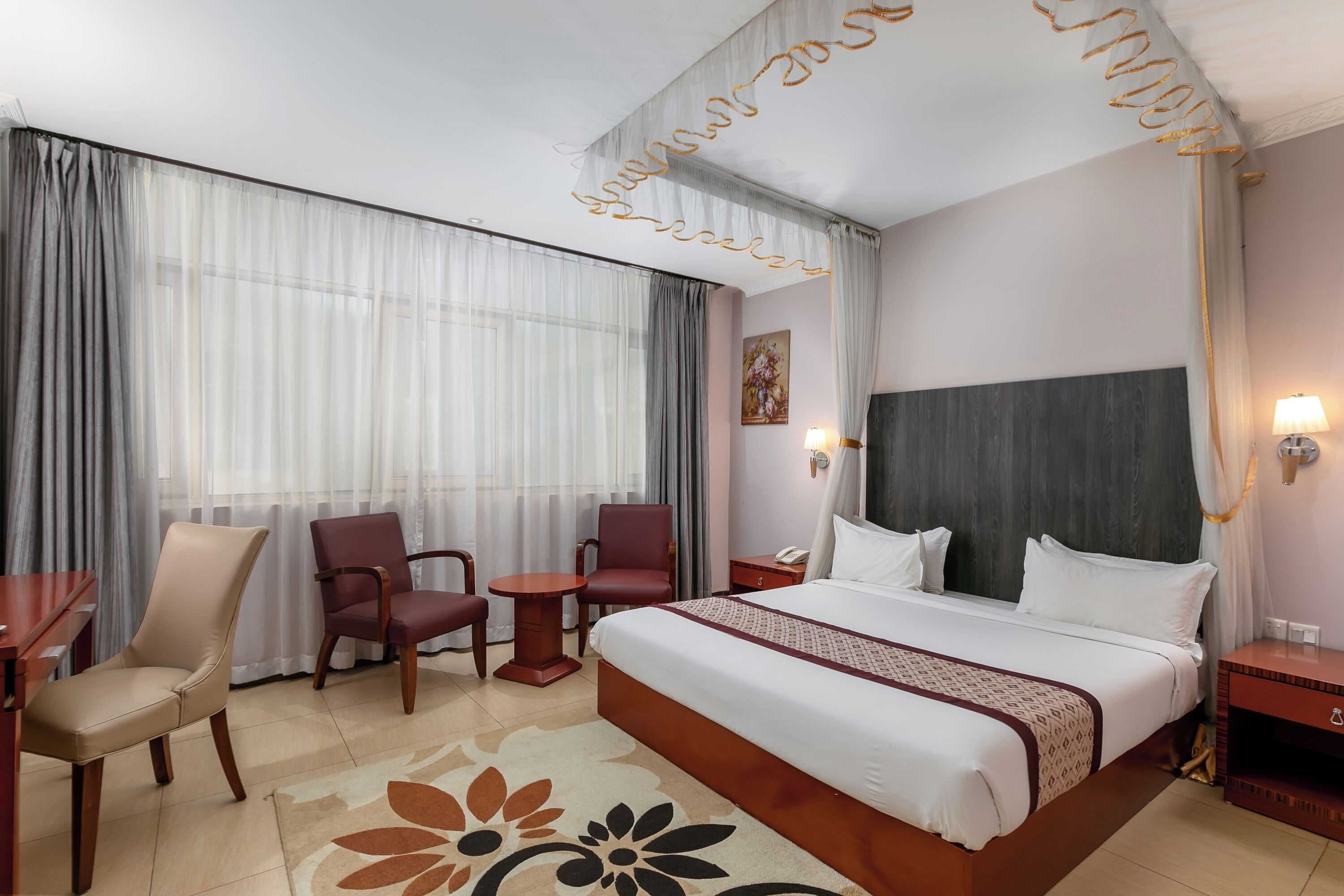 A hotel room with a large bed, two chairs, a small table, a nightstand, and a decorative wall hanging.