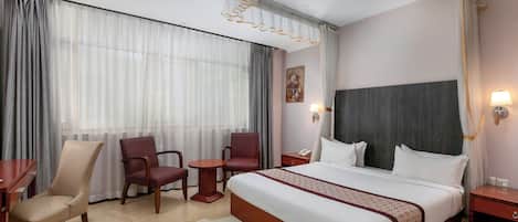 Habitación de hotel con una cama grande, dos sillas, una mesita pequeña, una mesita de noche y un adorno colgante en la pared.