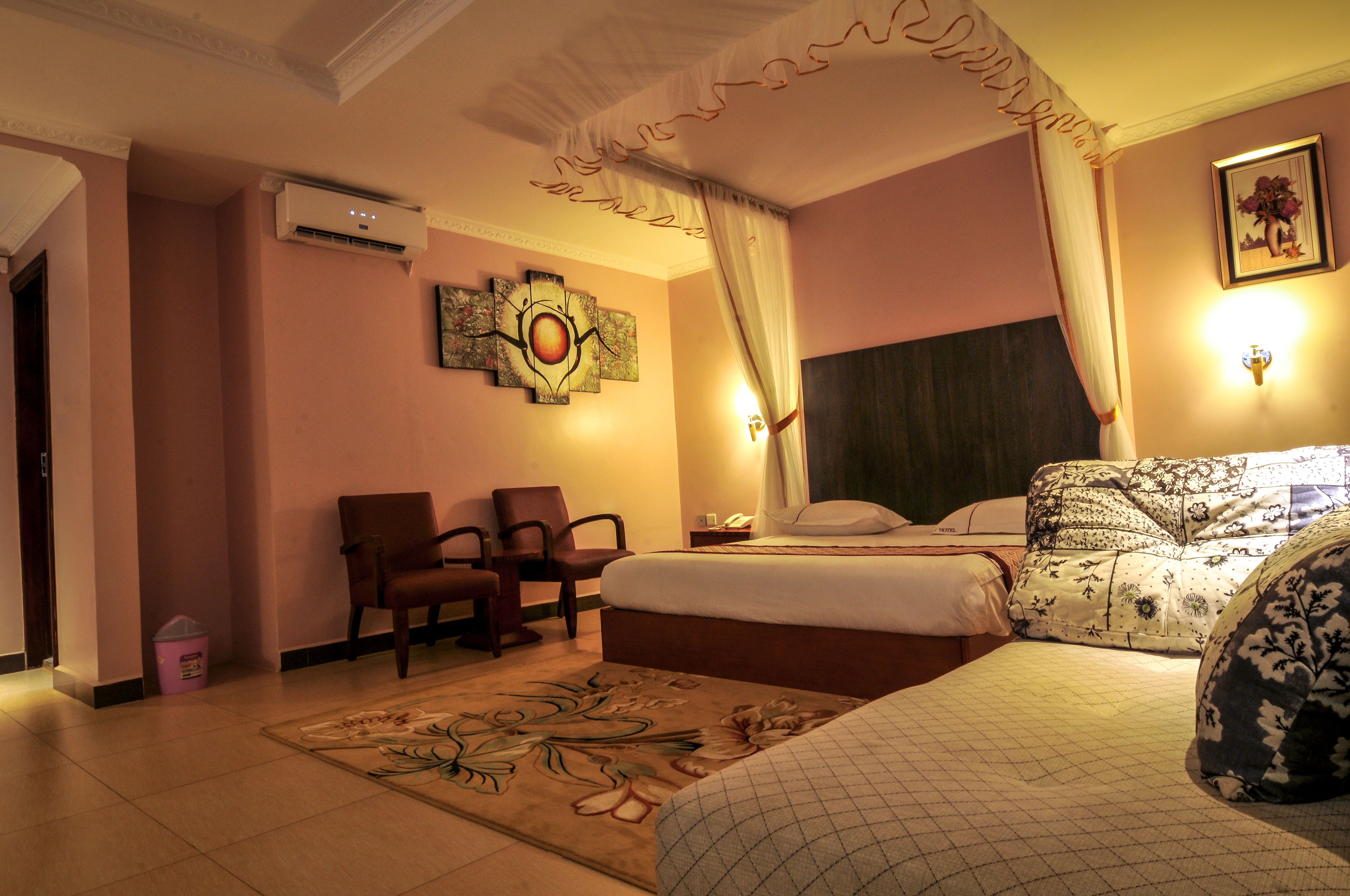 A hotel room with a bed, two chairs, a nightstand, an air conditioning unit, a decorative wall piece, and a framed picture.