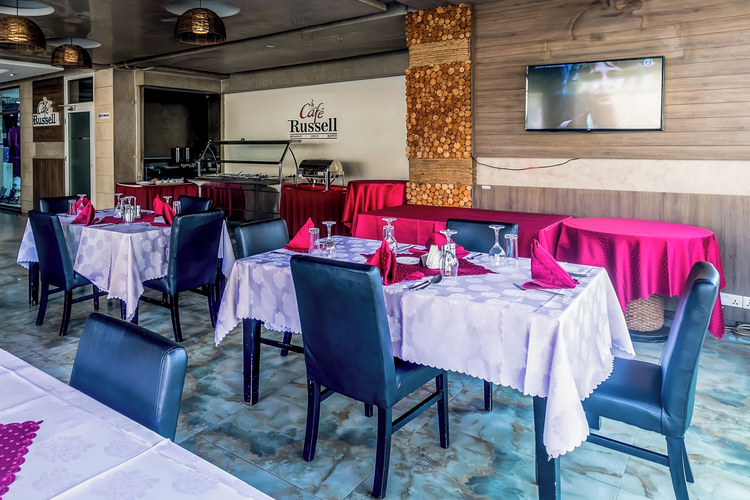 Un interior de restaurante con mesas dispuestas para comer, una zona de cocina y un televisor montado.