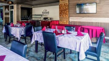 Interior de restaurante com mesas postas para refeições, área de cozinha e uma televisão na parede.