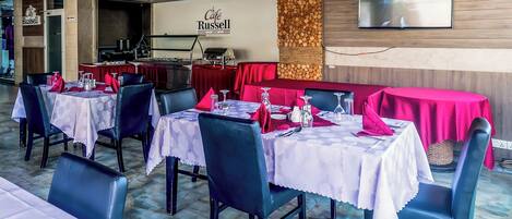 Un interior de restaurante con mesas dispuestas para comer, una zona de cocina y un televisor montado.