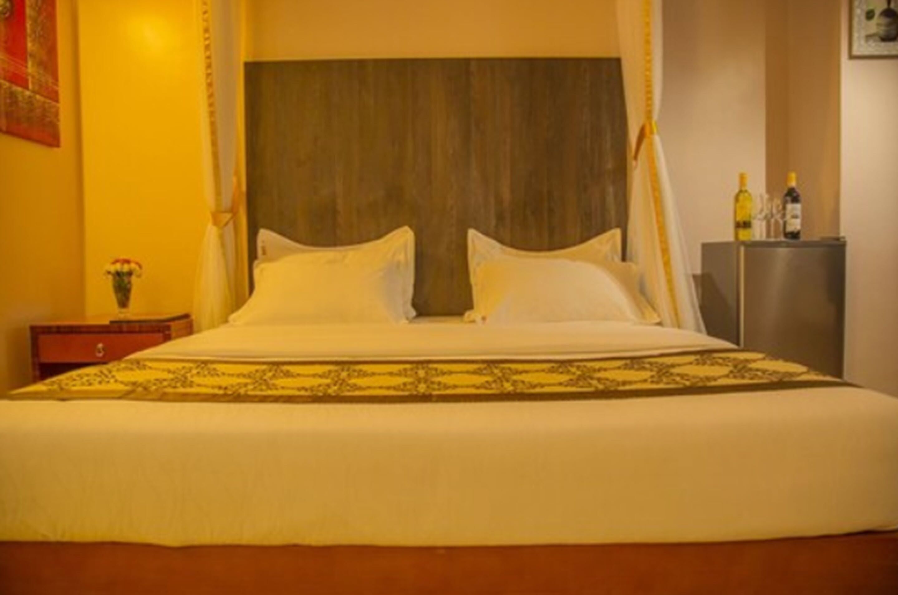 A large bed with a wooden headboard, a bedside table with a vase of flowers, and a minibar with bottles and glasses.