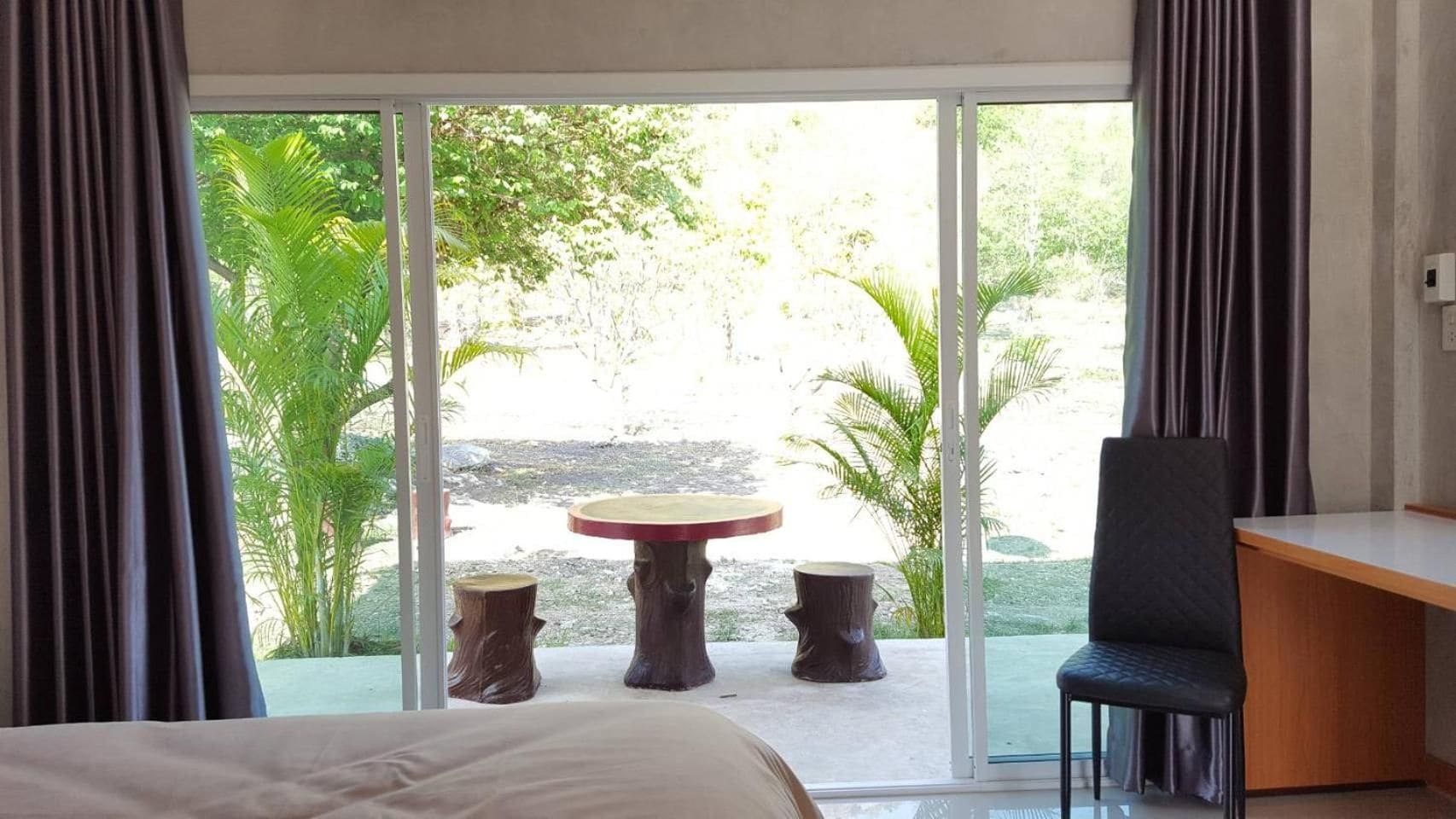 A hotel room with a bed, a desk, and a table with chairs. There is a sliding glass door leading to an outdoor area with trees and a view of the landscape.