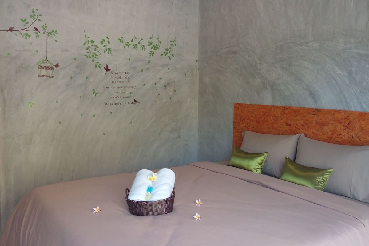 Un lit bien fait, avec un panier contenant des articles de toilette, un oreiller orné d’un motif floral et un mur décoré d’une fresque sur le thème de la nature.