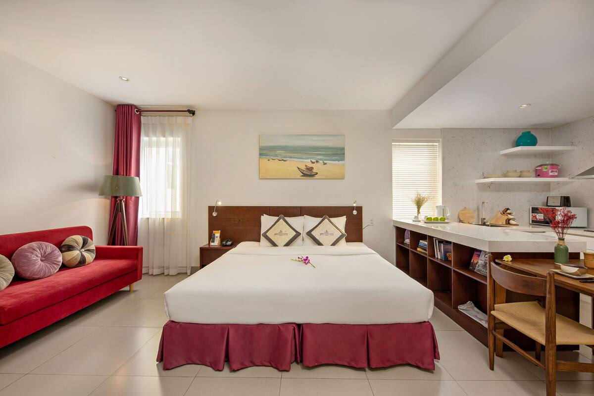 Habitación de hotel con una cama grande, un sofá rojo, una mesita de noche con lámpara, un cuadro con una escena de playa y una zona de kitchenette.