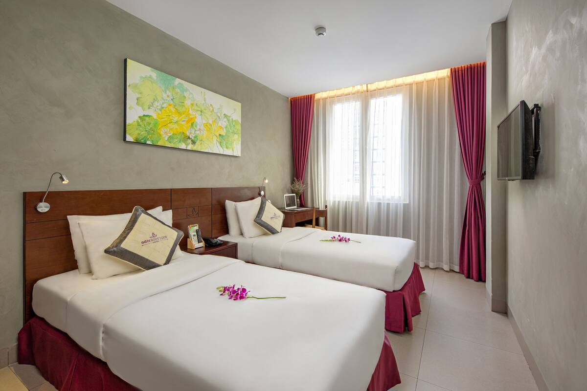 Habitación de hotel con dos camas, un cuadro en la pared, un televisor montado y una ventana con cortinas.