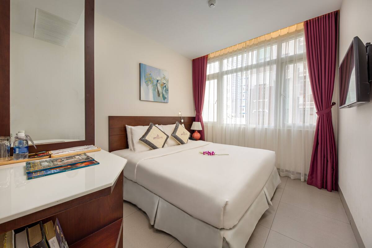 Habitación de hotel con una cama, un escritorio con lámpara, un cuadro en la pared y una ventana con cortinas.