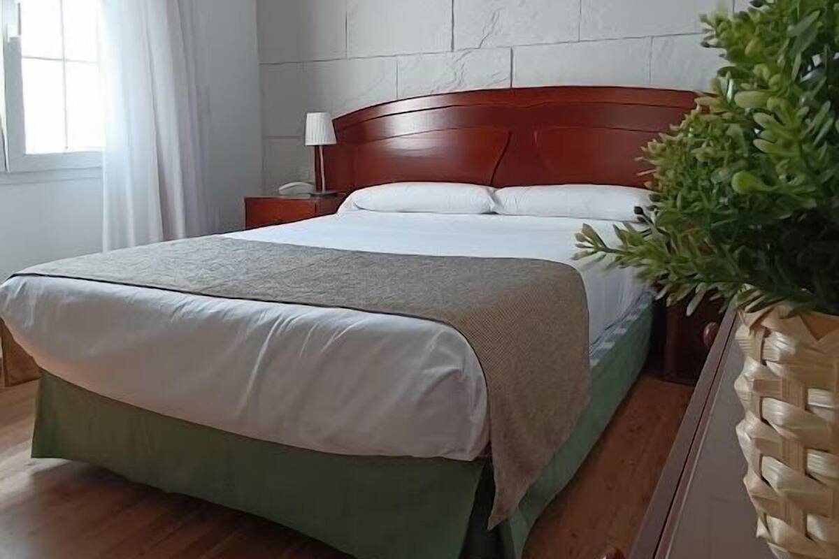 A bedroom with a double bed, a nightstand, a lamp, a potted plant, and a wooden dresser.