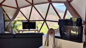 Una habitación moderna y bien iluminada, con un ventanal grande, un televisor de pantalla plana, una cafetera y un sofá negro.