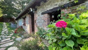 Un edificio rústico de piedra con fachada de ladrillo, un sendero de piedra y abundante vegetación, incluyendo flores coloridas.