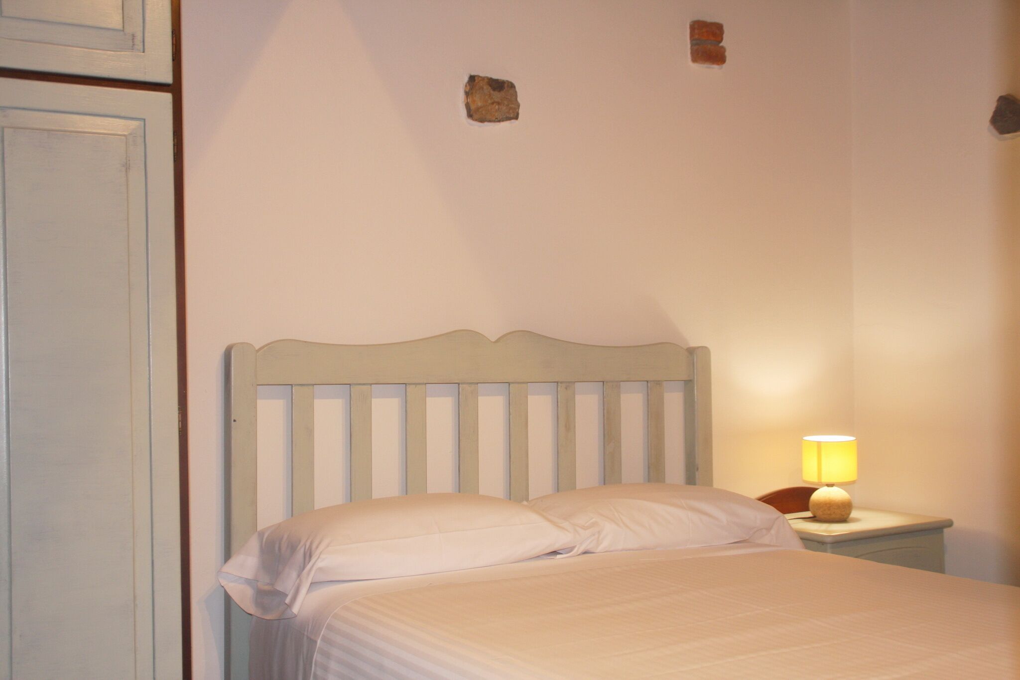 Ein Einzelbett mit Holz-Kopfstück, ein Nachttisch mit Lampe und eine schlichte Wand mit einem kleinen Bereich freigelegter Ziegelsteine.