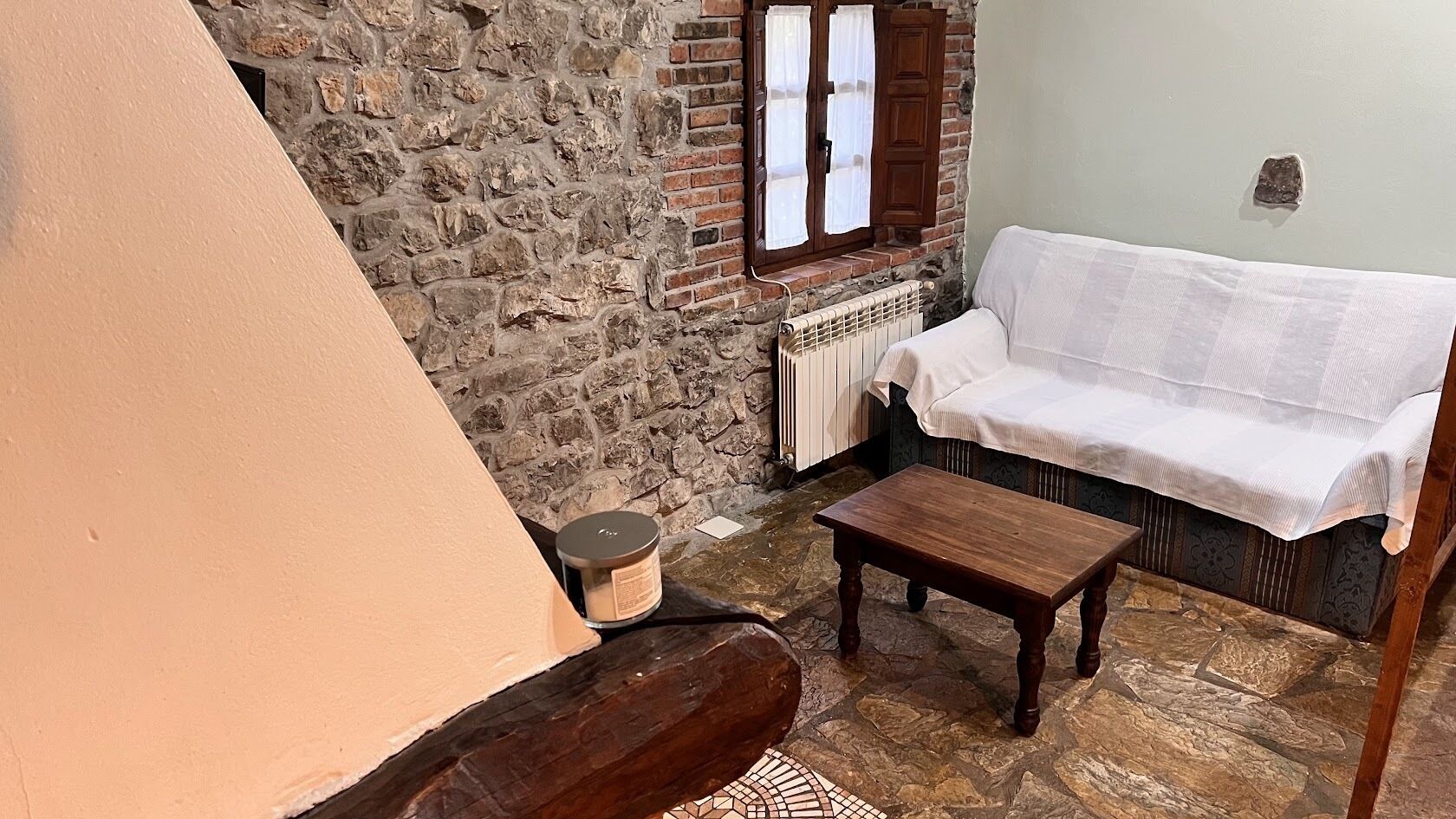 A cozy room with a stone wall, a white sofa, a wooden table, and a small candle on the floor.