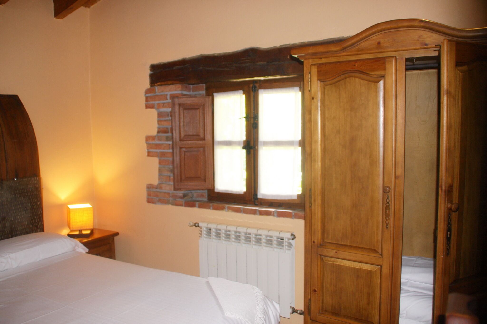 Een slaapkamer met een bed, een nachtkastje met een lamp, een houten garderobe, een bakstenen muur en een raam met luiken.