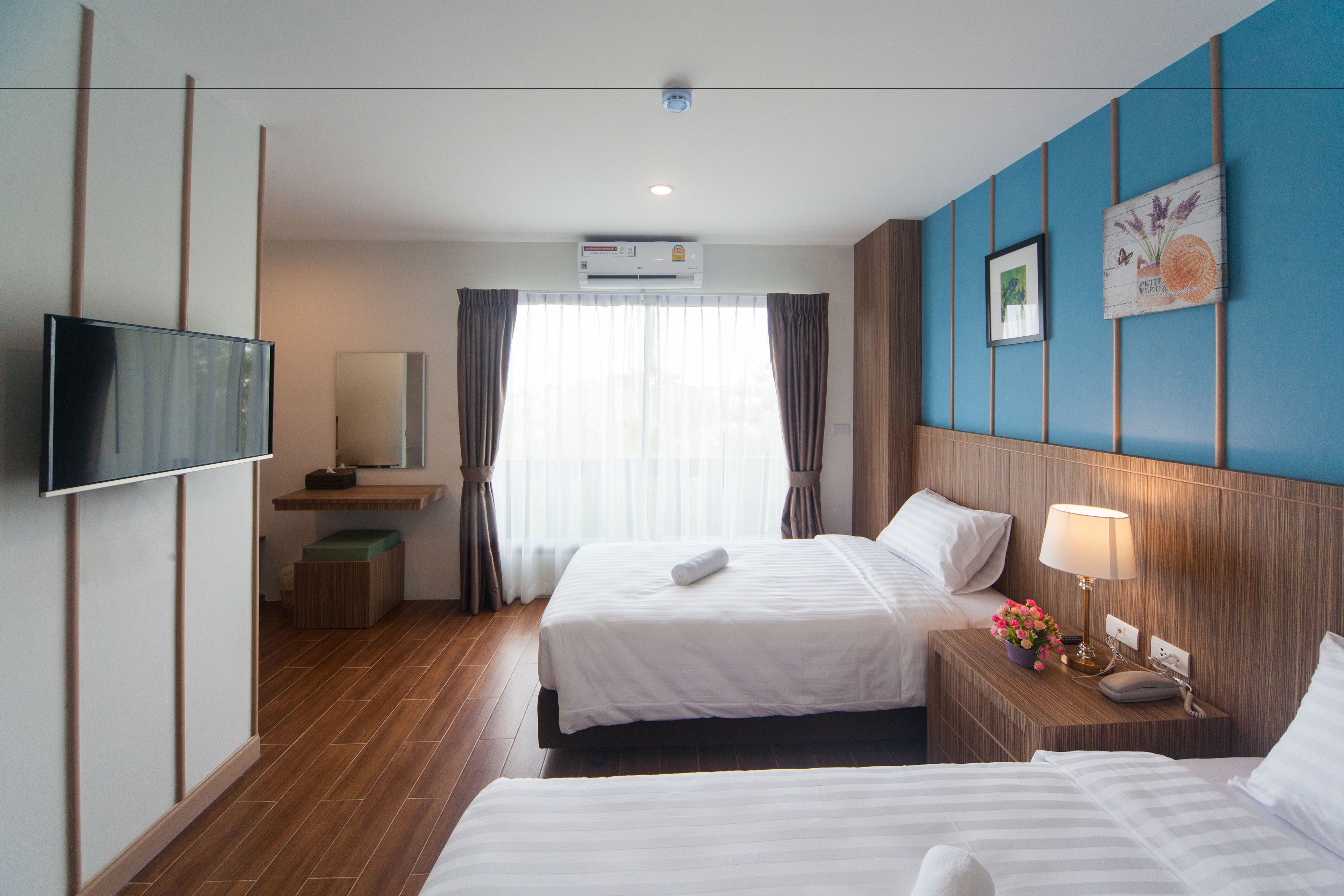 A hotel room with two beds, a flat-screen TV, an air conditioning unit, a bedside table with a lamp and flowers, and a large window with curtains.