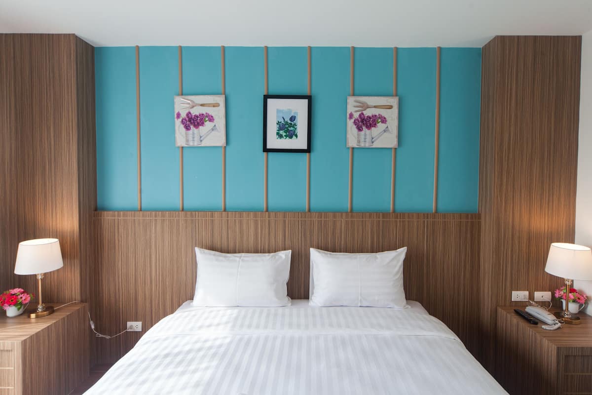 Una camera d'albergo con una testiera in legno, una parete decorata in tonalità verde acqua, due quadri, biancheria da letto bianca e abat-jour con fiori.