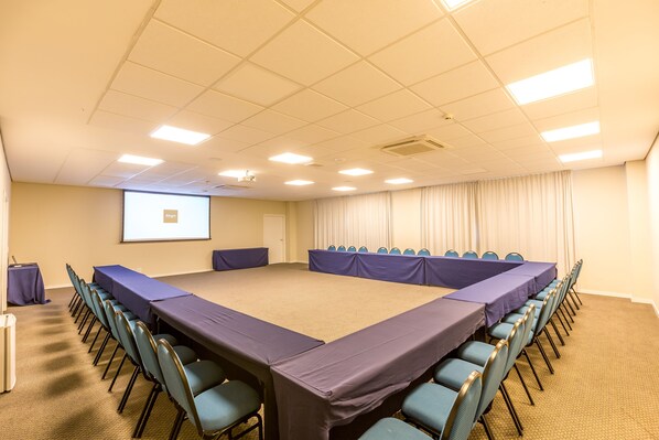 A conference room with a long table, chairs, a projector screen, and curtains. - Alegro Hotel (Jarinu)