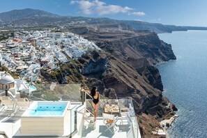 Une femme se tient sur un balcon qui donne sur une ville côtière, avec ses bâtiments blancs, une piscine et une vue dégagée sur la mer.
