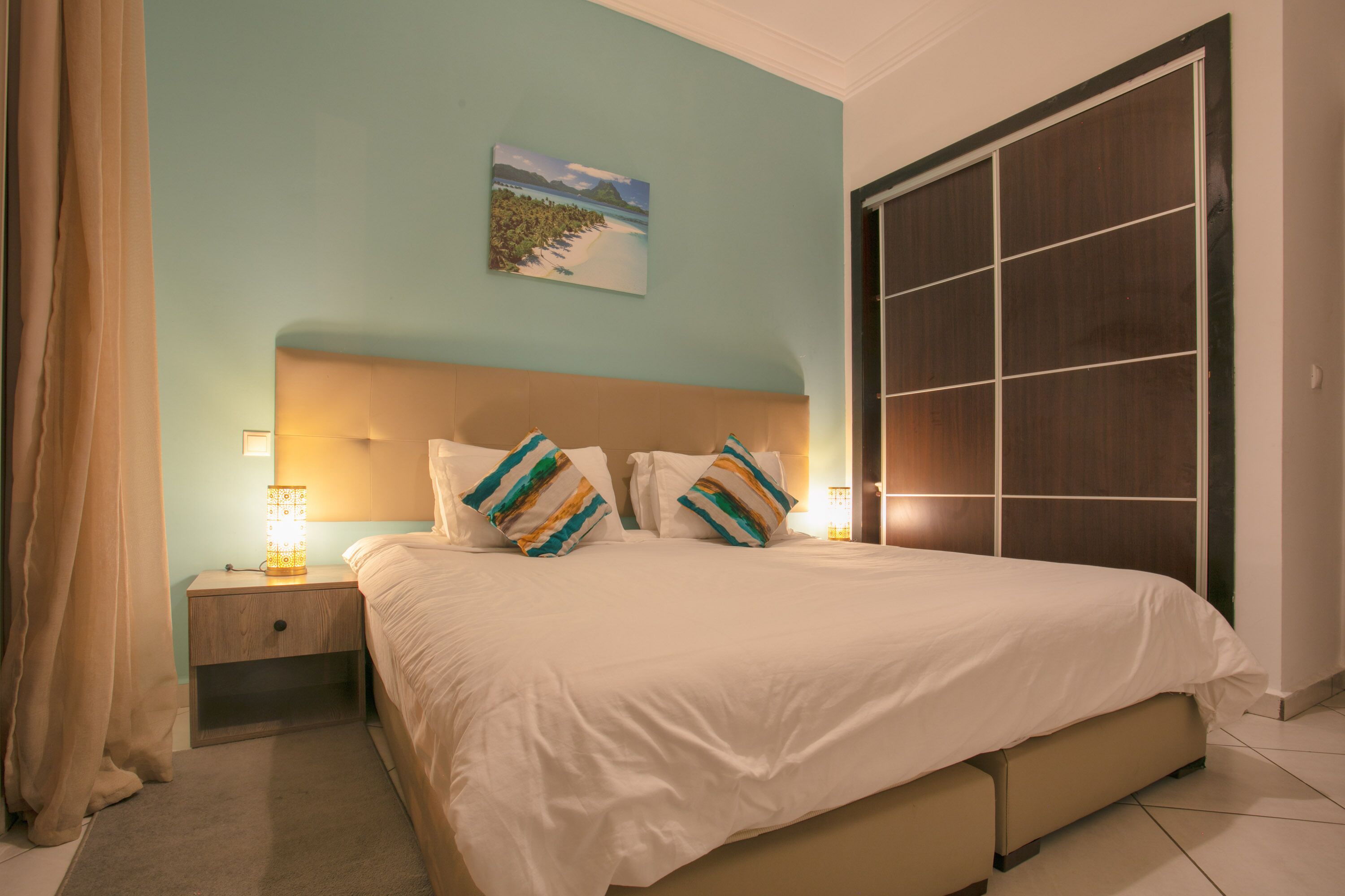 A neatly made bed with a headboard, a bedside table with a lamp, and a painting on the wall.