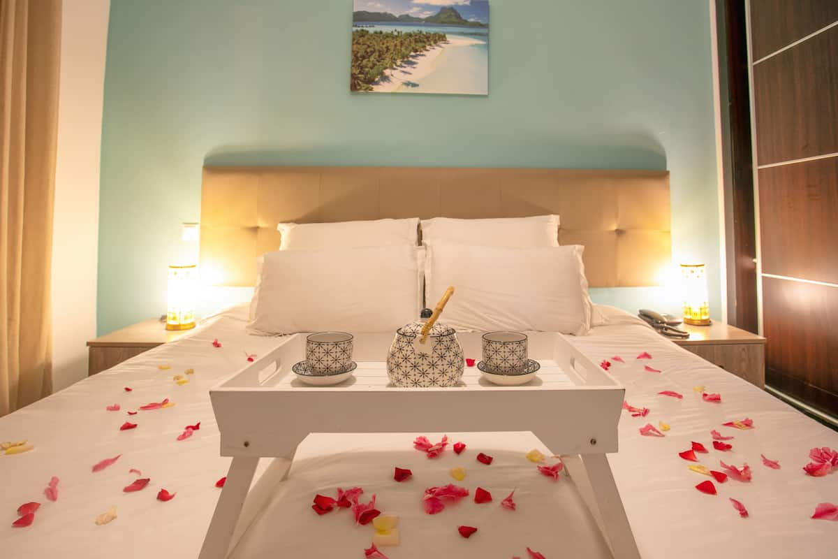 Un lit parfaitement fait, avec du linge de lit blanc et des pétales de rose éparpillés sur la couverture. Sur le lit, on aperçoit un plateau avec deux tasses et une théière. La pièce est décorée d'un mur bleu clair et d'un tableau représentant une scène de plage, placé au-dessus du lit.