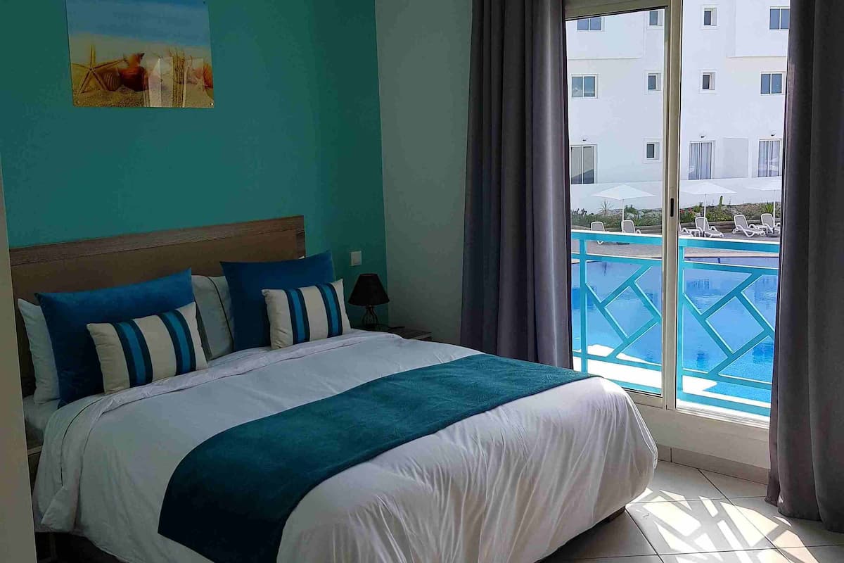 Une chambre d’hôtel avec un lit, un tableau au mur, une fenêtre donnant sur une piscine et des bâtiments, et un luminaire au plafond.