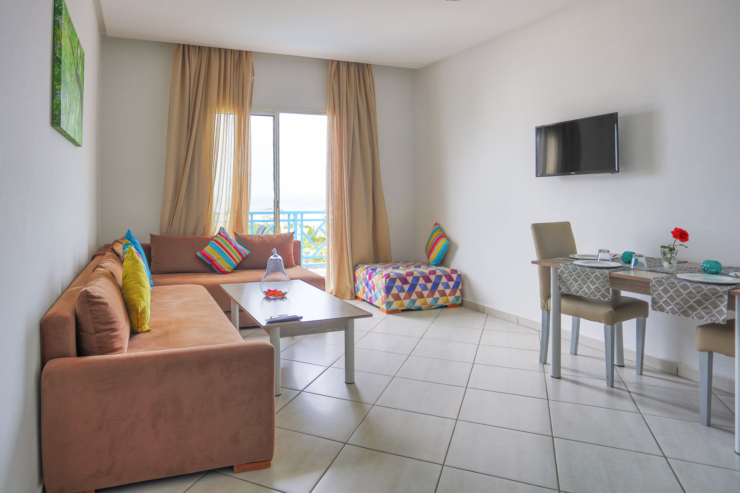 A modern living room with a sofa, a coffee table, a dining table with chairs, a TV mounted on the wall, and a balcony with curtains.