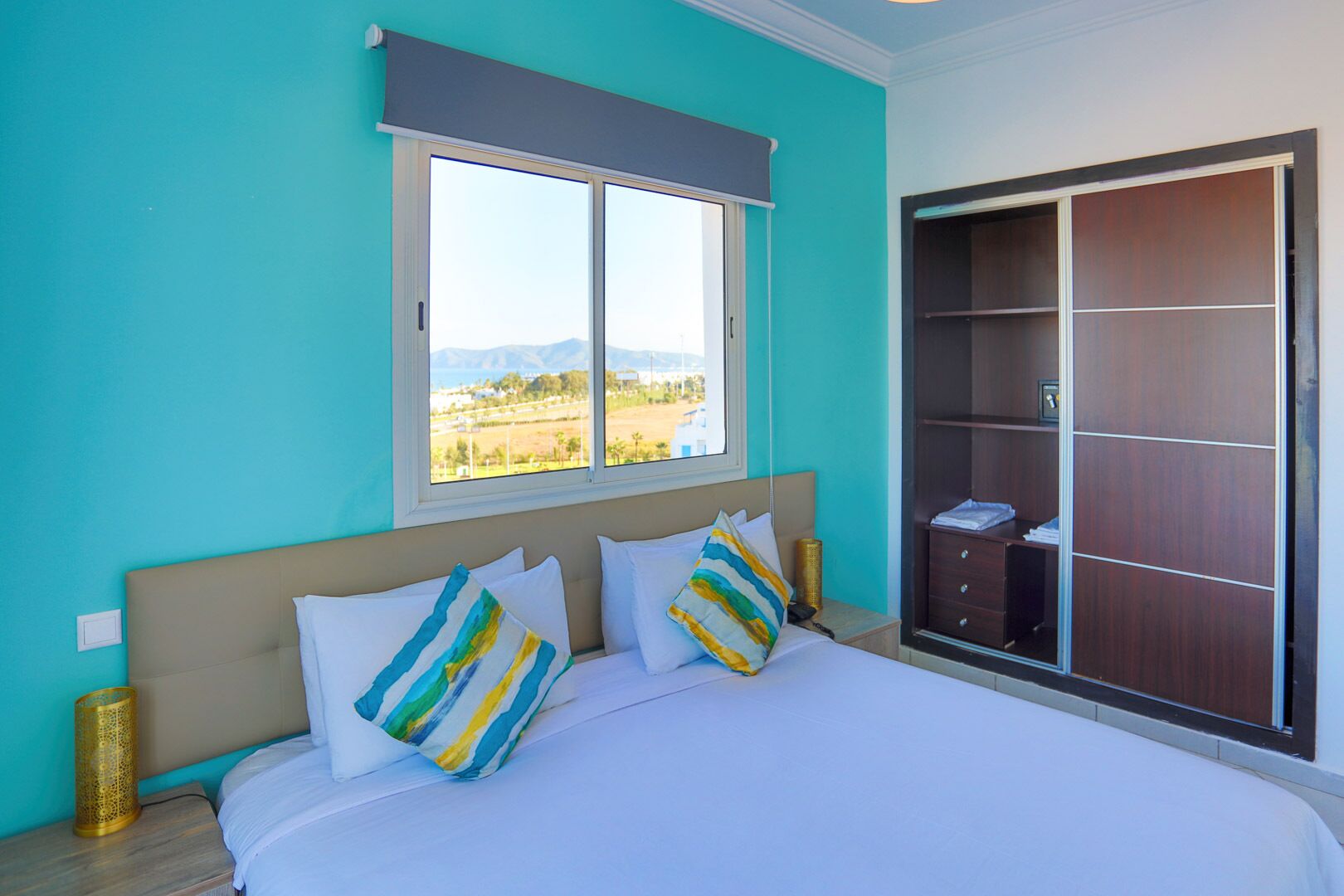 Un dormitorio con cama, dos almohadas, una mesita de noche, una lámpara, una ventana con vistas y un armario.