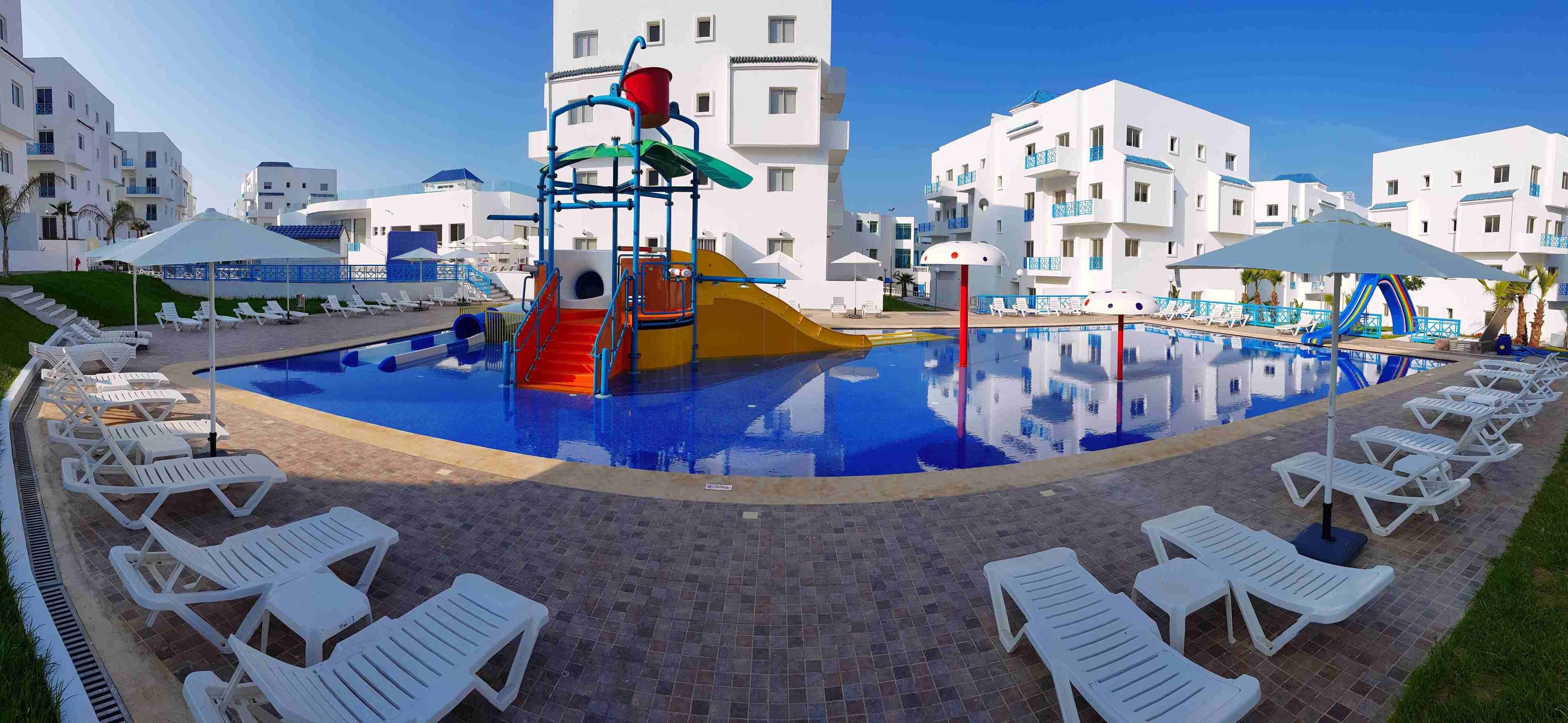 A swimming pool with a slide and a play structure, surrounded by white lounge chairs and a poolside area with umbrellas.