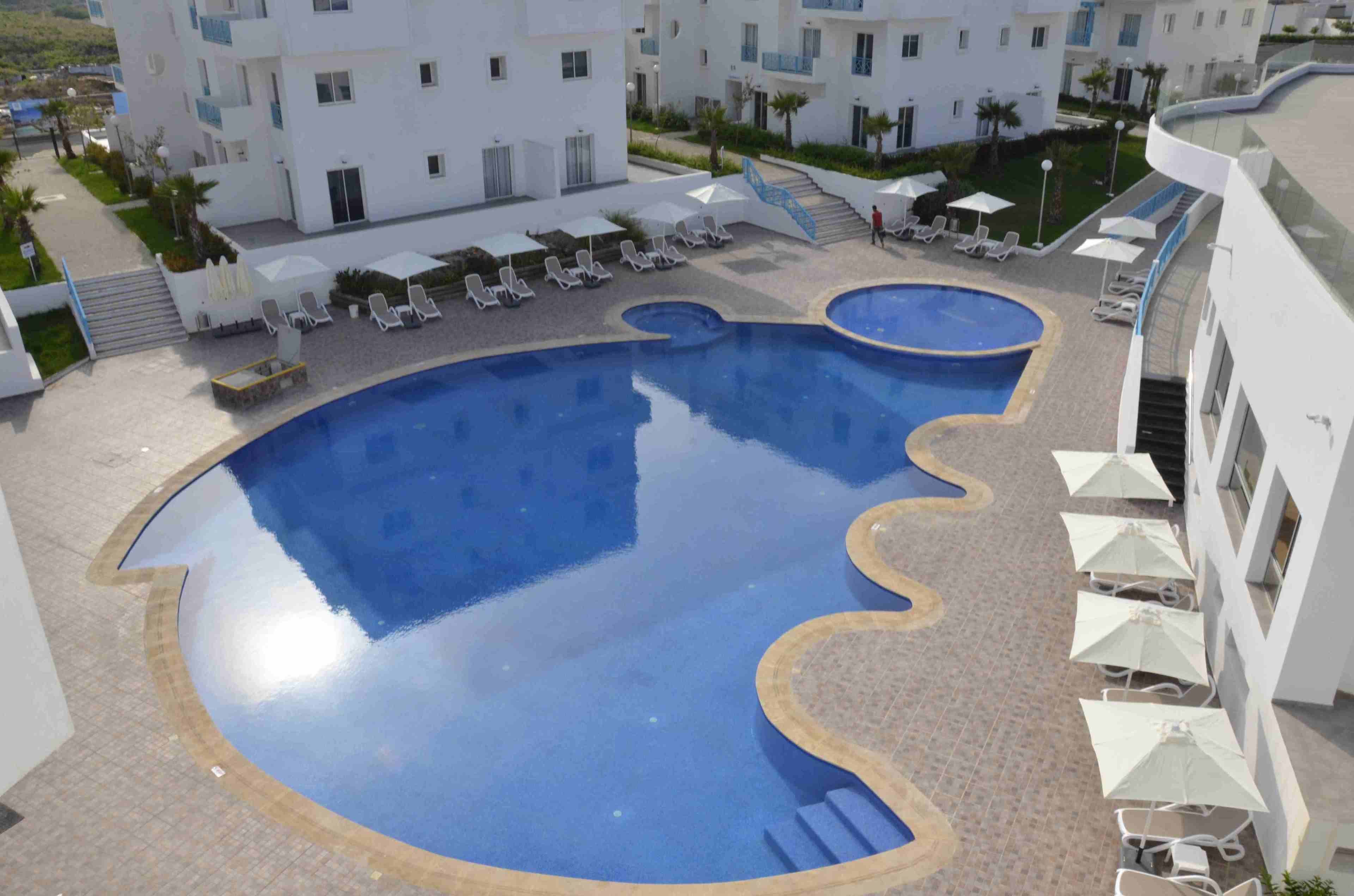 En pool med en vågformad kant, omgiven av solstolar och parasoller, med vita byggnader intill och en trappa som leder upp till en övre nivå.