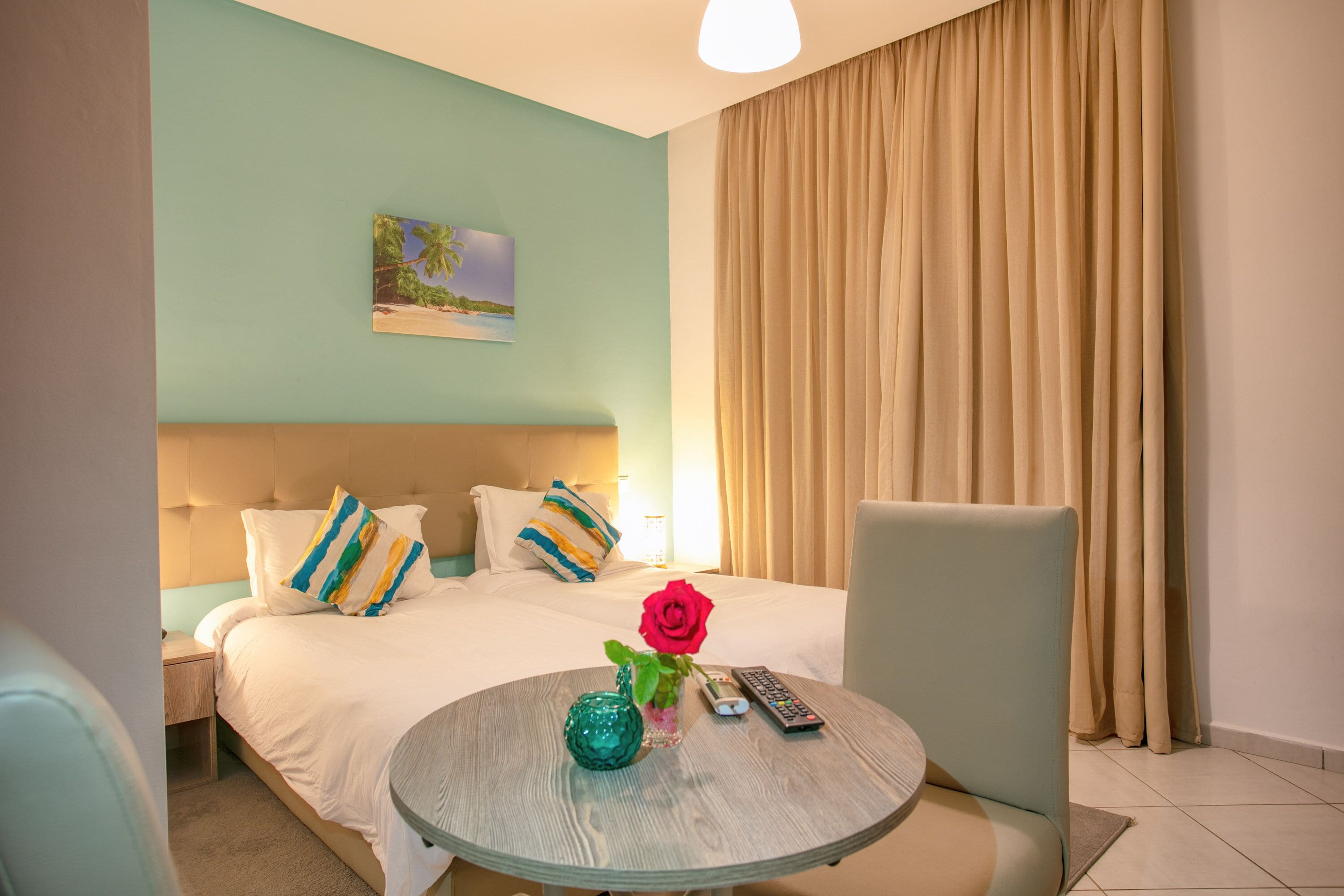 A hotel room with a bed, a round table with a vase and a remote control, and a painting on the wall.
